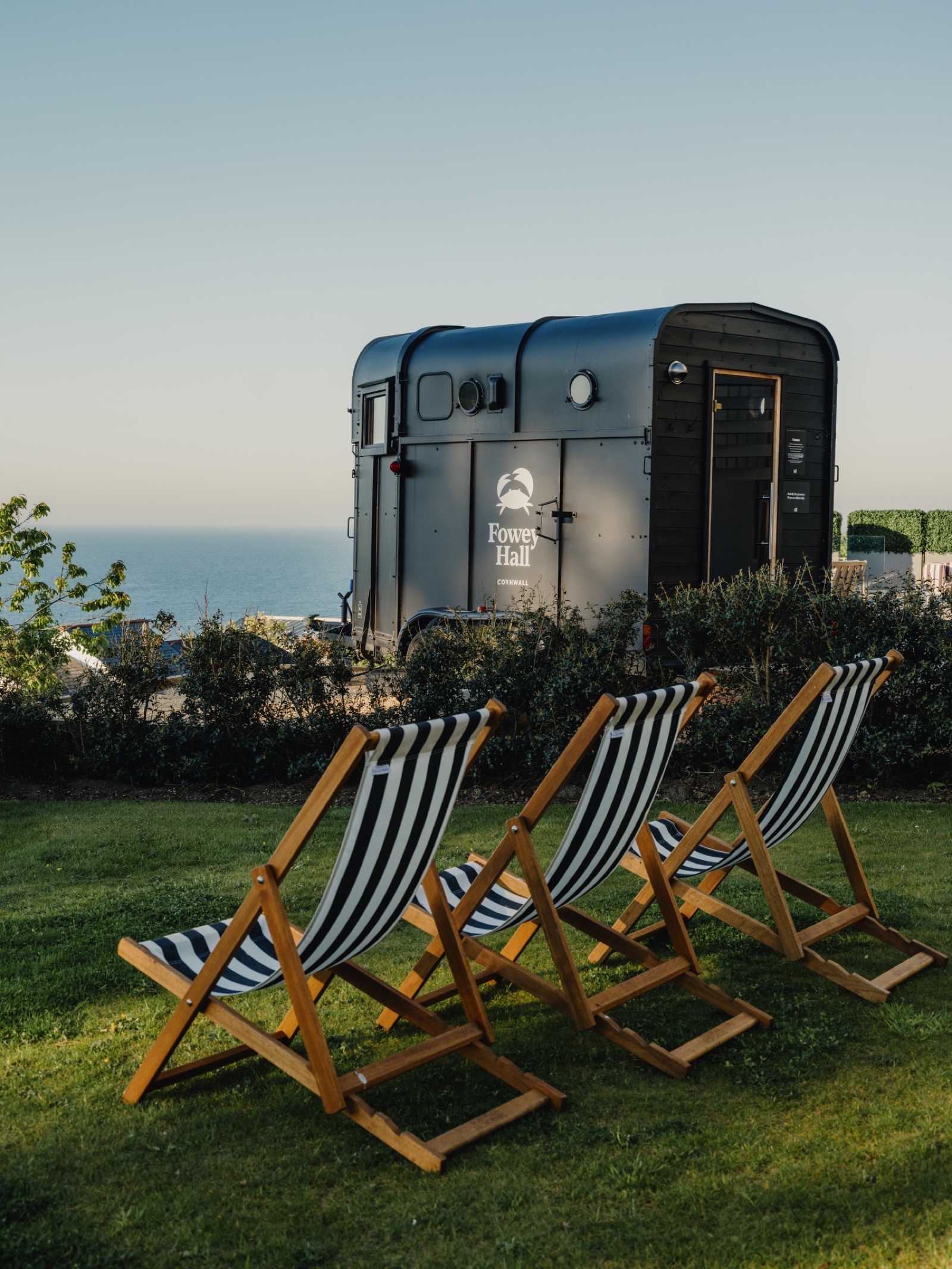 Three striped deckchairs on the Fowey Hall lawn facing the black horsebox sauna above the sea