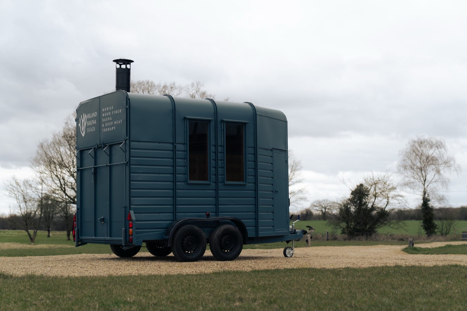 Island Sauna Shack's navy horsebox sauna in a Cambridgeshire field, chimney and branded livery visible from the rear three-quarter angle