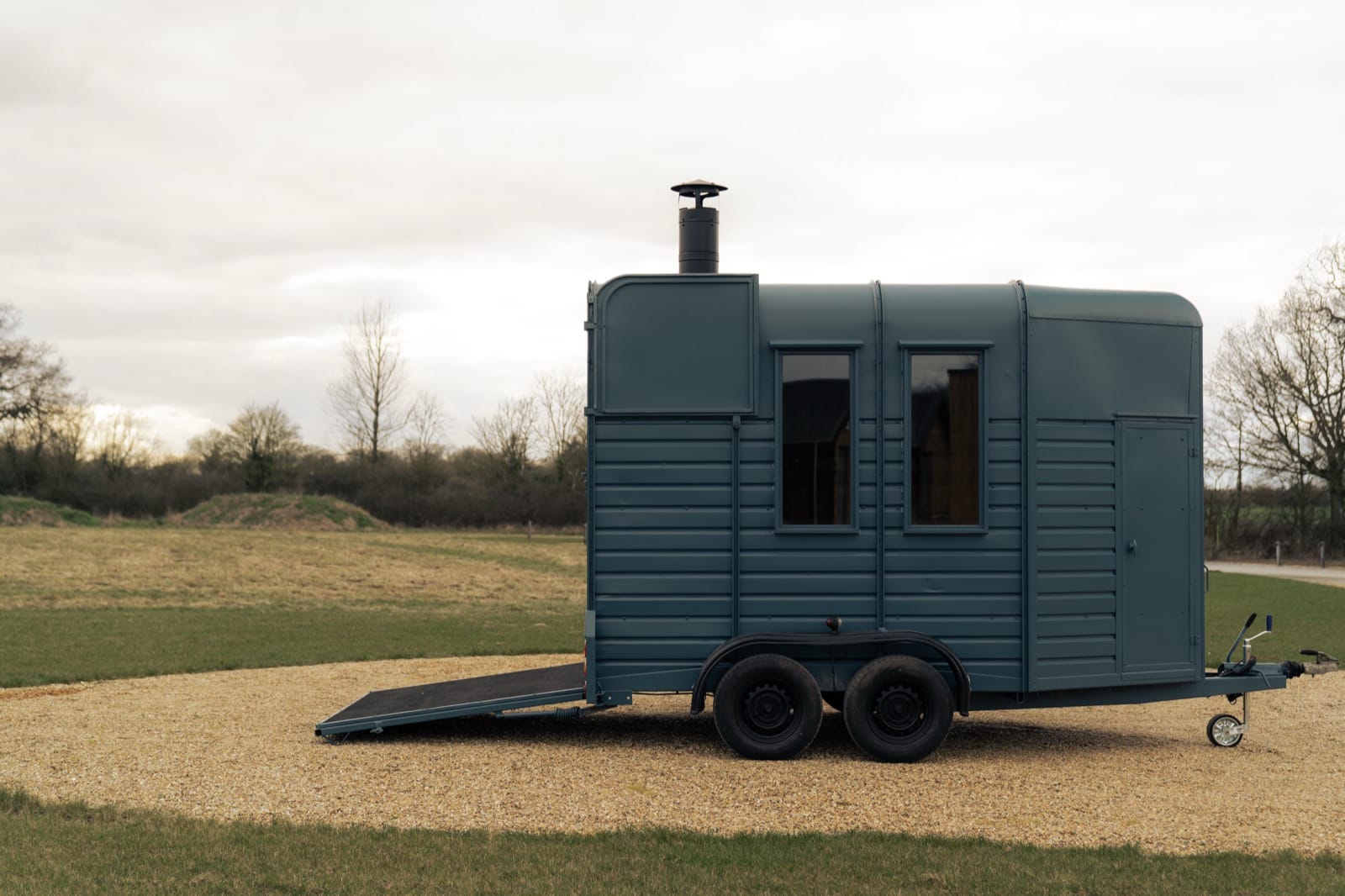 Side profile of the Island Sauna Shack horsebox with its chimney stack, parked on gravel with open fields behind