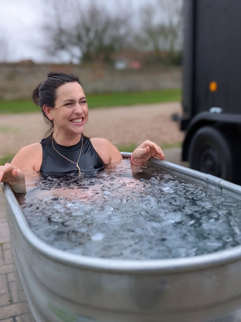 A woman laughing in an ice bath, a black horsebox sauna behind her