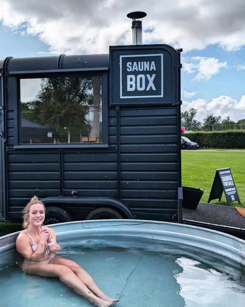 A young woman relaxing in a plunge tub in front of a black Sauna Box horsebox sauna