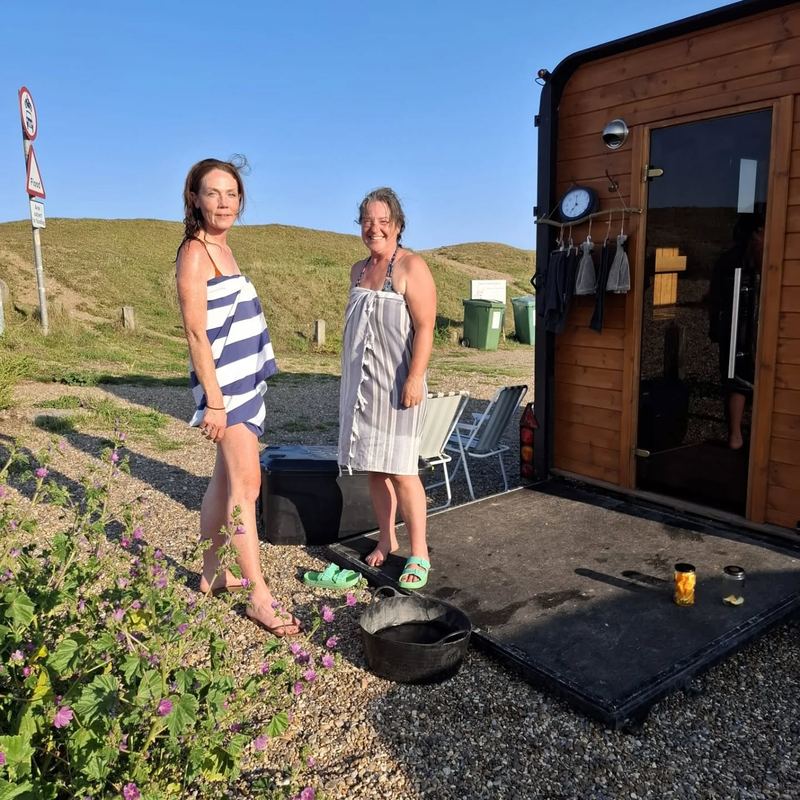 Two women in striped towels at the door of a wooden horsebox sauna in the dunes