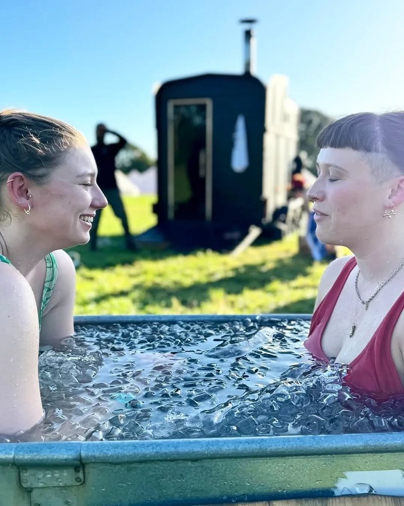 Two women chatting in an ice bath, a sauna on the grass behind them