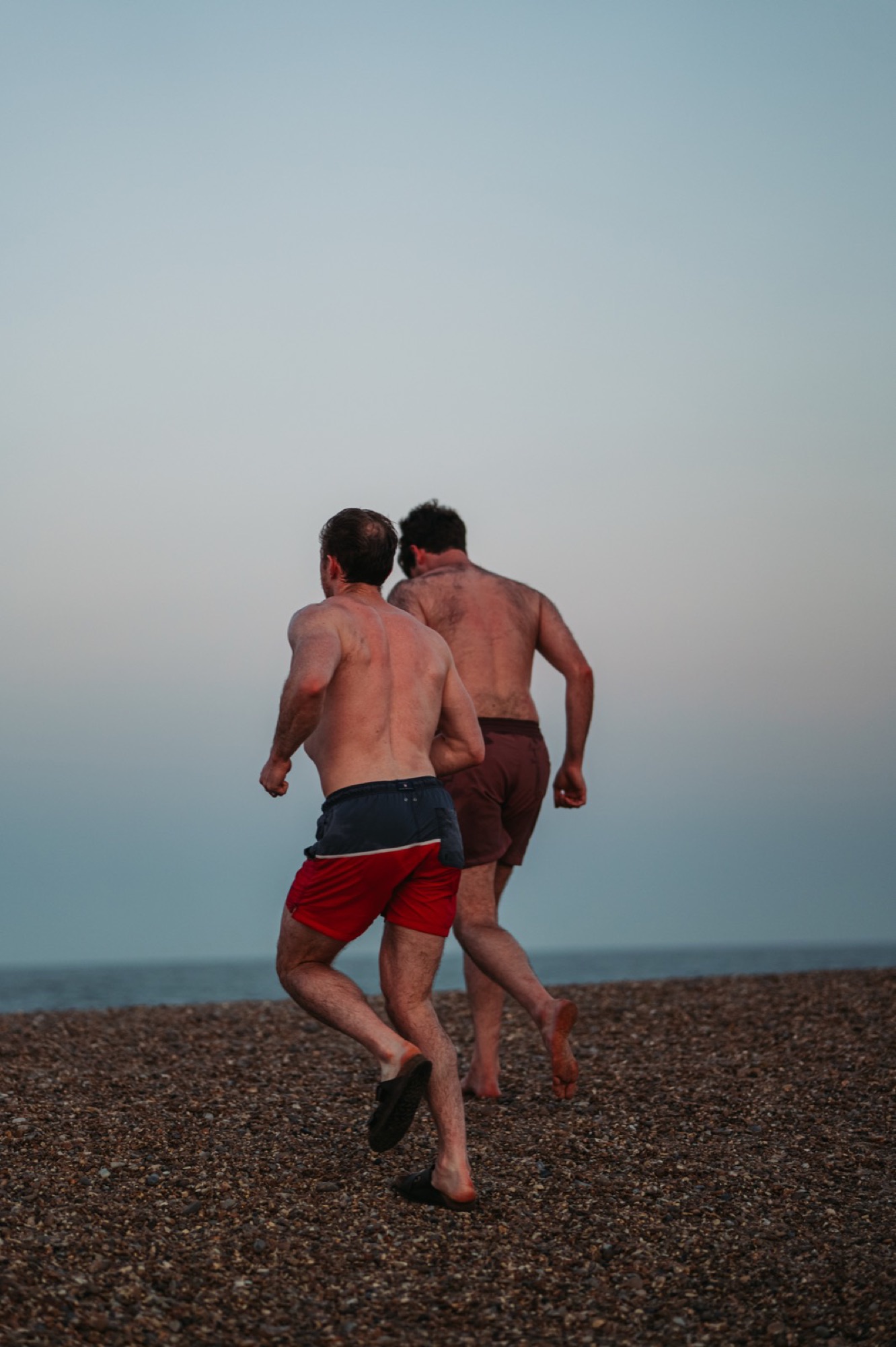 Two men run toward the sea across a pebble beach at dusk for a cold dip